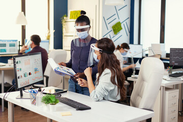 Obraz premium Multiethnic coworkers at workplace analysing marketing statistics wearing face mask against covid19. Diverse team working respecting social distancing during global pandemic with coronavirus. New