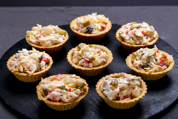Tartlets with salad on a black wooden round board laid out in a circle on a gray concrete background.