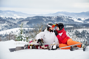 Charming young woman looking at man and smiling while wrapping arm around his shoulder. Joyful boyfriend and girlfriend sitting on snow-covered hill in mountains. Concept of skiing and relationships.