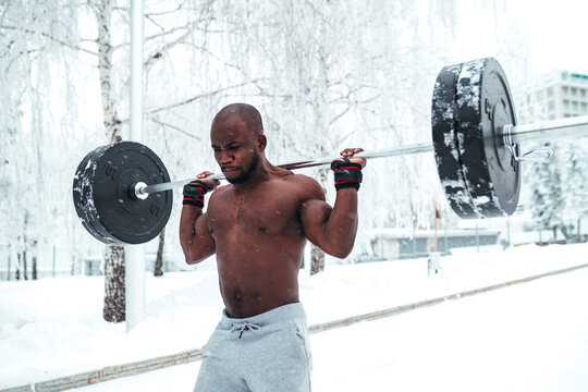 A guy with a naked torso does exercises with a barbell on a background of snowy trees in winter