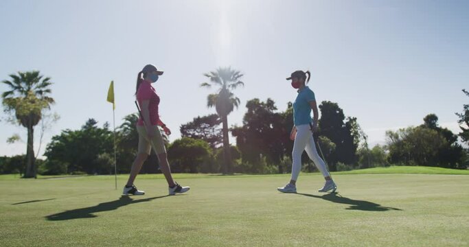 Two caucasian women playing golf wearing face masks greeting each other with feet