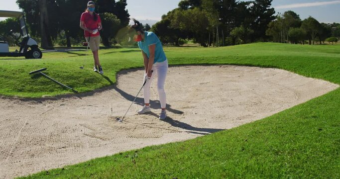 Two caucasian women playing golf wearing face masks one taking shot from bunker