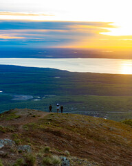 Obraz premium Three people stand atop a mountain overlooking a small town, an inlet, and a sunset.