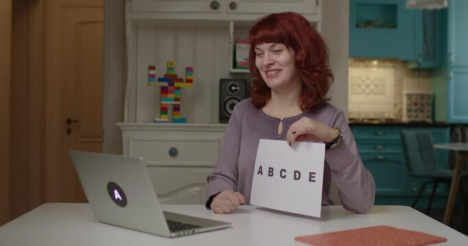 40s female teacher talking to laptop and holding sheet of paper with ABCDE letters. Online education and distance learning process. Work from home English school teacher.