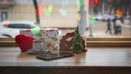 A heart made of red thread, a cup of coffee and a gift box, a smartphone with a toy Christmas tree with a bokeh effect, on the background of the window and highlights in the form of hearts. Happy Vale
