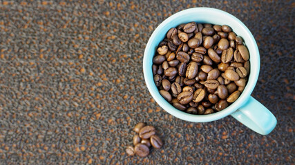 roasted dark brown coffee beans are poured into a turquoise mug with a handle and several coffee beans next to it on a black textured background top view