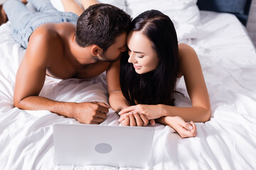 smiling sexy couple holding hands near laptop on bed on blurred background