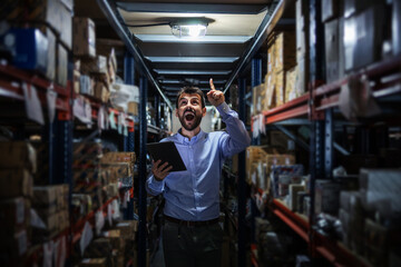 Obraz na płótnie Canvas Three quarter length of smiling bearded businessman standing in warehouse full of goods for delivering and holding tablet. He is having great idea.