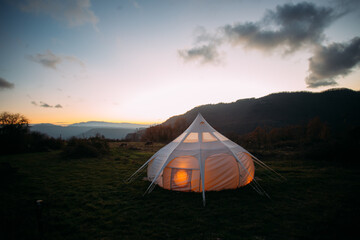 Night time sunset shot of glamping camping tent set up in beautiful nature landscape. Cosy and warm big tent lit from inside with orange dimmed lights. Romantic weekend getaway for couple © BublikHaus