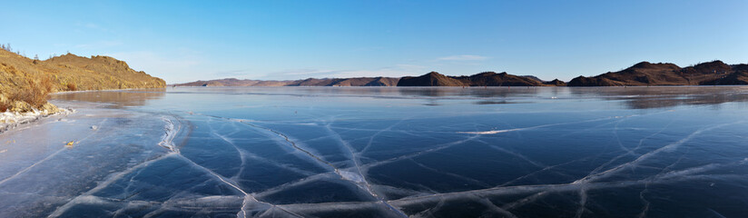 Frozen Baikal Lake on a sunny winter day. Panoramic view of beautiful blue smooth ice with cracks in Kurkut Bay. Tourists walking and skating on ice are visible in the distance. Natural background