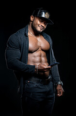 Fototapeta na wymiar Strong athlete in dark jeans poses to the camera on black background. Unbuttoned black shirt and perfect abs. Portrait of a afro american bodybuilder with phone in hands.