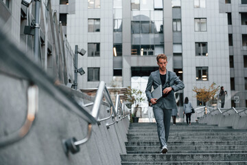 Laptop in hands. Young elegant man in good clothes is outdoors in the city at daytime