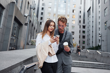 Fototapeta premium With phone and drink. Woman and man in the town at daytime. Well dressed people