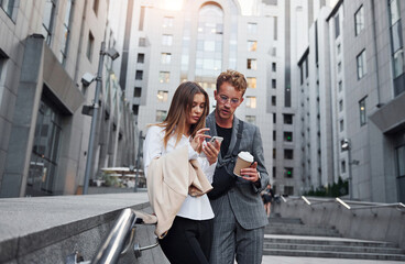 Fototapeta premium With phone and drink. Woman and man in the town at daytime. Well dressed people
