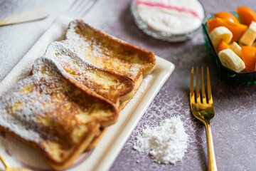 Traditional french toast with cinnamon on a gray background. Traditional Spanish sweet fried toasts, torrijas, with banana and persimmon, fresh cream, powdered sugar on a ceramic plate, and gold