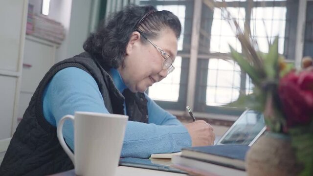 Senior asian woman wear glasses doing work from home by tablet online cooking class lesson, sit write with pencil on notebook, cold winter scene, distanced leaner, happy retirement housewife activity