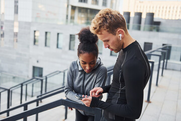 Using phone. European man and african american woman in sportive clothes have workout together