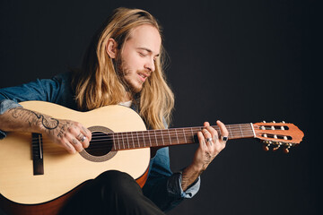 Portrait of long haired tattooed musician looking dreamy playing on acoustic guitar composing new song  over black background - obrazy, fototapety, plakaty