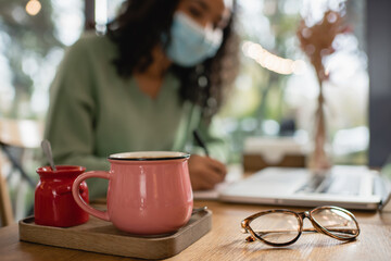cup of coffee near sugar bowl and african american woman in medical mask on blurred background