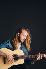 Obraz premium Side view of long haired tattooed musician looking sensual playing on acoustic guitar over black background. Stylish singer with guitar