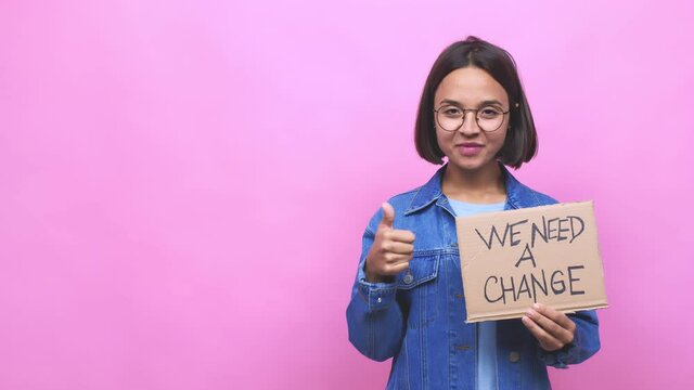 Young hispanic woman holding a we need a change cardboard smiling and raising thumb up