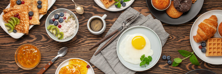 Continental breakfast captured from above (top view, flat lay). Coffee, tea, croissants, jam, egg, pancakes, maffins and oatmeal. Wooden background. Family breakfast table.
