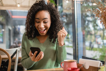 excited african american woman with clenched fist looking at smartphone in cafe