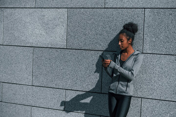 Uses phone. Young african american woman in sportive clothes have workout outdoors at daytime