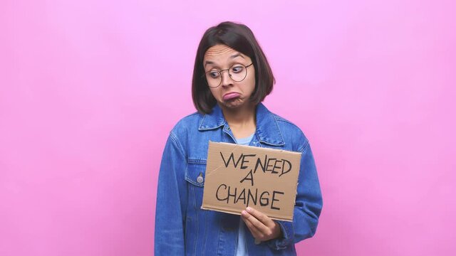 Young hispanic woman holding a we need a change cardboard shrugs shoulders and open eyes confused