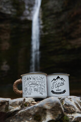 Two creative mugs and rest after hard day of hiking. Tea party in hike on background of waterfall. Interesting angle water from waterfall fills enameled white camping mug with inscription.
