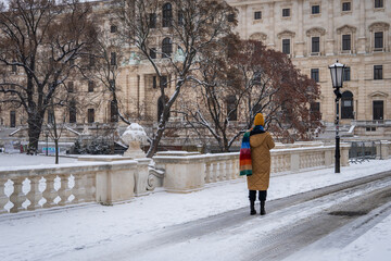 A woman dressed in colorful, stands out, winter clothes standing in an urban city park covered with white snow. Contrast colors of clean white and fun, vibrant, colors. 