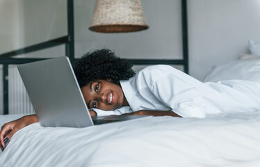 Lying down on bed with laptop. Young african american woman with curly hair indoors at home