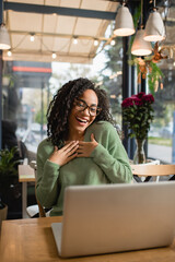 flattered african american woman in glasses looking at laptop on blurred foreground