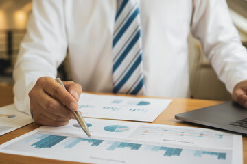 Asian finance staff is sitting at the table. The hand is holding a pen pointing to a graph document showing the company's growing profits, financial business ideas. Corporate profit growth