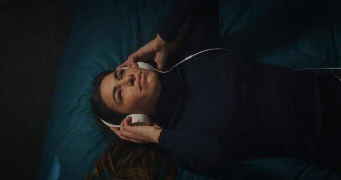 Cinematic shot of young woman is putting on headphones and relaxing her mind by listening her favorite music playlist or audio book while lying on a bed in bedroom at home with soft atmospheric light