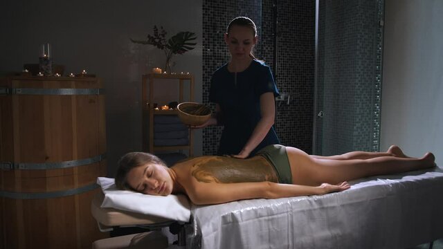 Wrapping the body of a young woman with seaweed and mud in a spa salon.