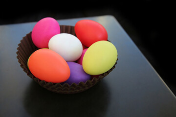 Multi colored painted easter eggs in red, pink, yellow, green and white in a brown paper basket against a dark background.