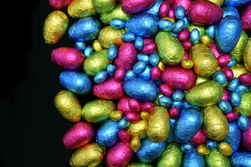 Fototapeta na wymiar Pile or group of multi colored and different sizes of colourful foil wrapped chocolate easter eggs in pink, blue, yellow and lime green against a black background with copy space.
