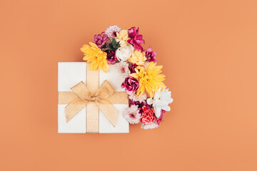 Obraz premium A mix of various flowers next to a beautiful white box wrapped with a ribbon, a beautiful bow on the lid of the box, a gift for March 8 on an orange background