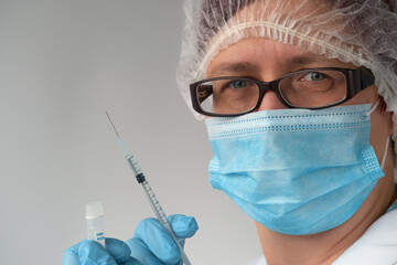 Coronavirus vaccine concept. Medic, doctor or nurse with covid-19 vaccine vial and syringe. Mature European female tech in face mask, glasses, disposable hat, blue gloves looks at camera.