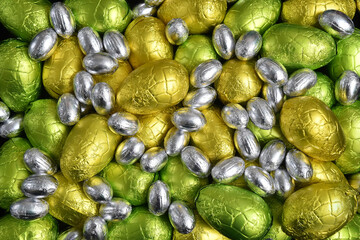 Large & small yellow, gold, green, lime green and silver spring colours of foil wrapped chocolate easter eggs, against a black background.