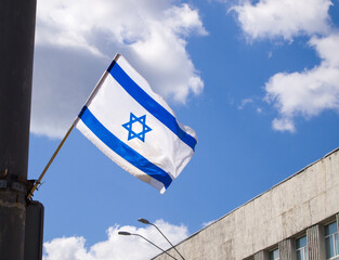 White and blue flag of Israel with a blue star of David in the middle over a bright sky with clouds.