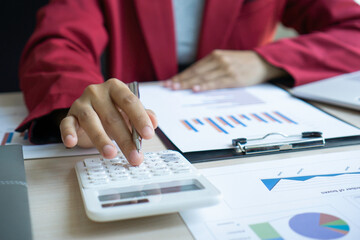 An Asian female finance worker is pressing on a calculator to calculate the company's earnings based on numbers and documents, bar graphs on her office desk, financial and profit report ideas.