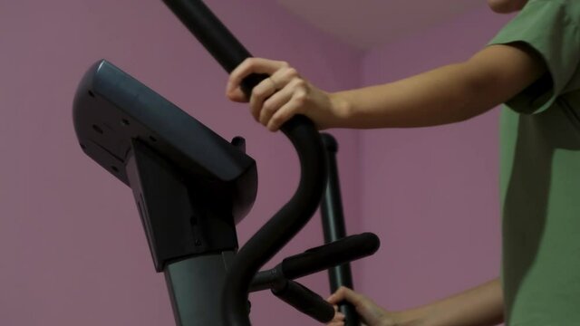 Beautiful athletic young girl makes settings on an elliptical trainer in the gym. Concept. Side view of a woman doing sport exercise on pinkk wall background.