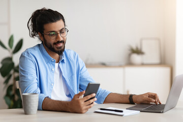 Fototapeta na wymiar Arab Freelancer Guy Using Smartphone While Working On Laptop In Home Office