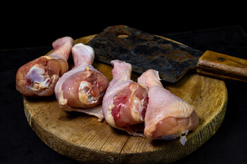 Raw chicken legs on a wooden board. Chicken legs on a black background, an old kitchen hatchet lies nearby. Chicken meat.