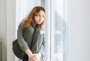 Beautiful sad unhappy teenager girl with curly hair sitting on the window sill