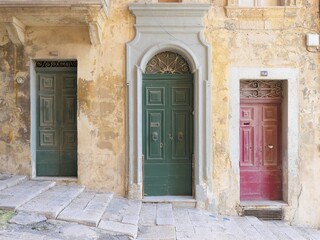 Valletta, Malta, December 25, 2020: Grey, green and red weathered wooden doors set into ochre-coloured sandstone wall on a slopped and stepped street of Malta.
