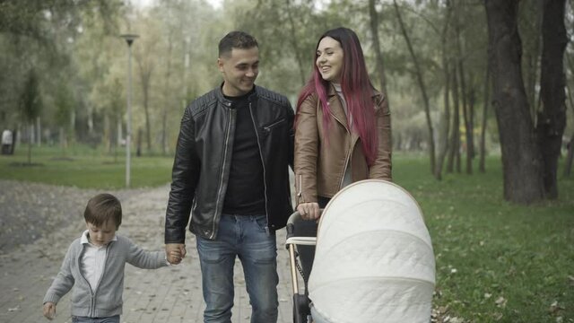 Portrait of happy young Caucasian man walking holding hand of little son and kissing woman pushing baby stroller. Cheerful family strolling in spring or autumn park outdoors. Unity and leisure concept