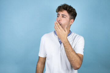 Young handsome man wearing a white t-shirt over blue background with his hand over his mouth and surprised, looking side